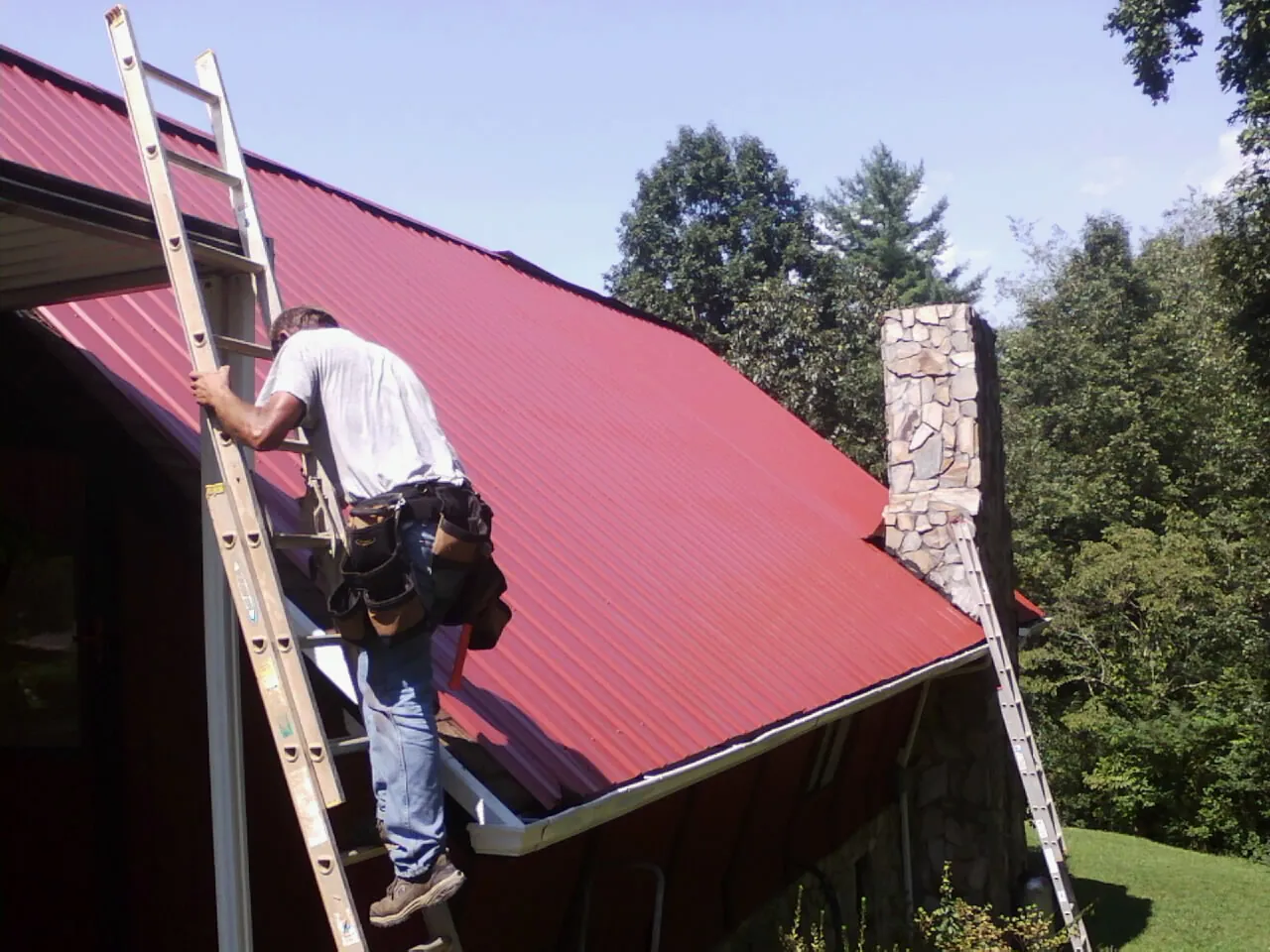 Roofer on a ladder beside a red metal roof and stone chimney — Soffit & Fascia Repair by Roofing Brothers in Amston